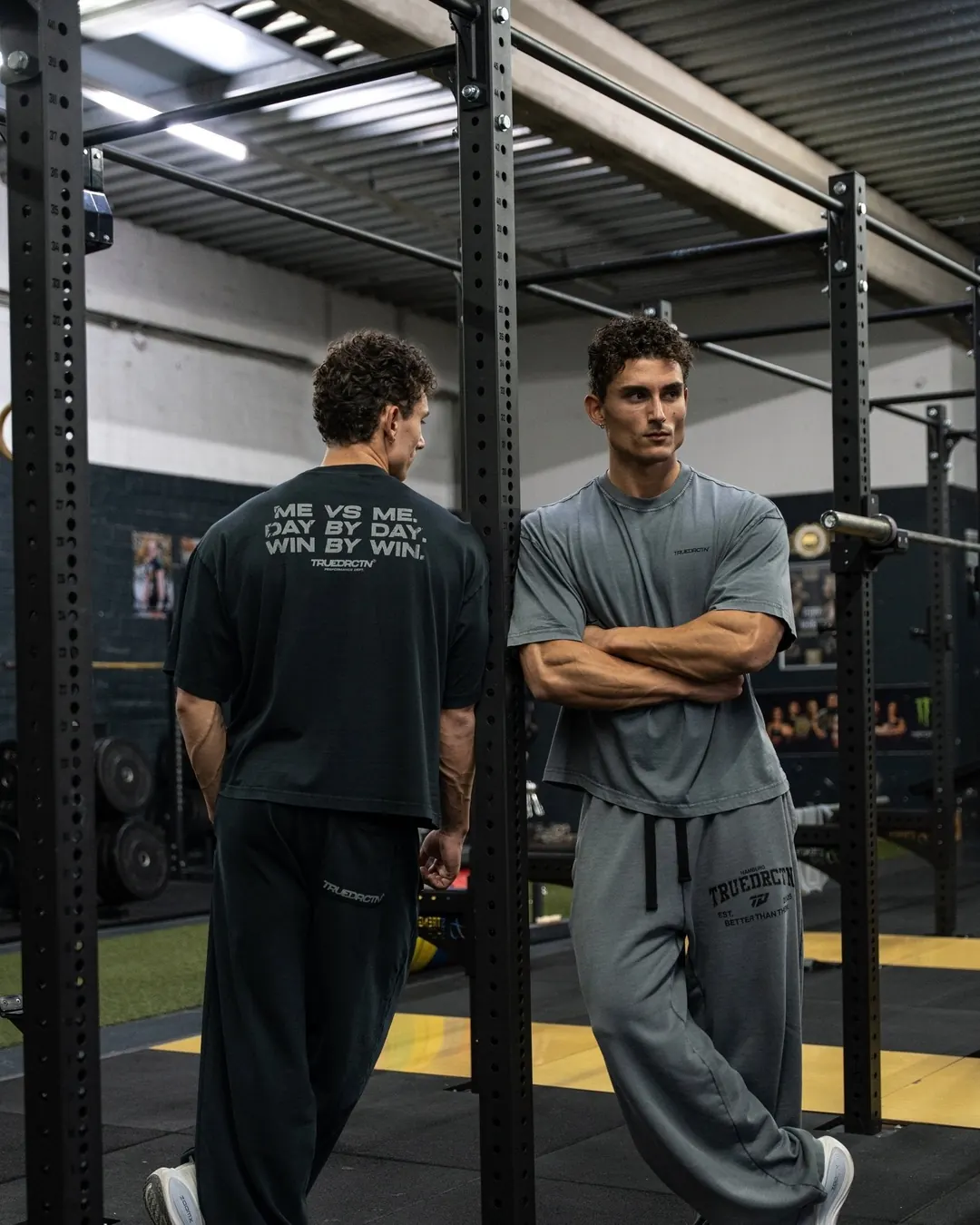 Zwei Athleten im Me vs Me Tee und im grauen Tee lehnen am Rack